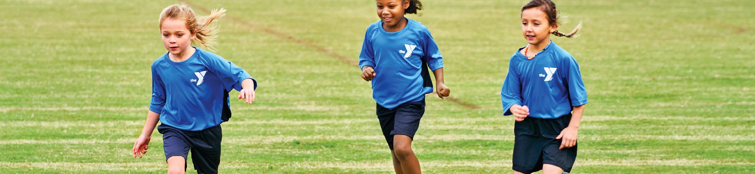 Three girls in blue soccer uniforms running towards a ball.